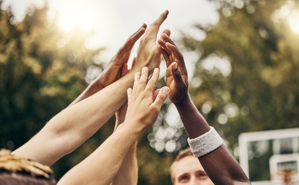 A group of diverse hands reaching up and touching together in a high five outdoors, symbolizing teamwork and unity, with blurred trees and a basketball hoop in the background.