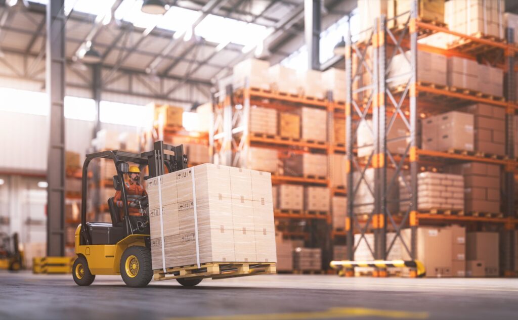 A worker drives a yellow forklift carrying a large pallet of boxes in a spacious warehouse filled with shelves stacked with packages and cartons. Sunlight shines through high windows, illuminating the organized storage area.