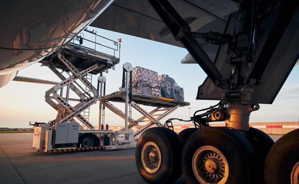 A cargo loader lifts a large, net-covered pallet to the open hold of an airplane on an airport tarmac, with the plane’s landing gear visible in the foreground at sunset.