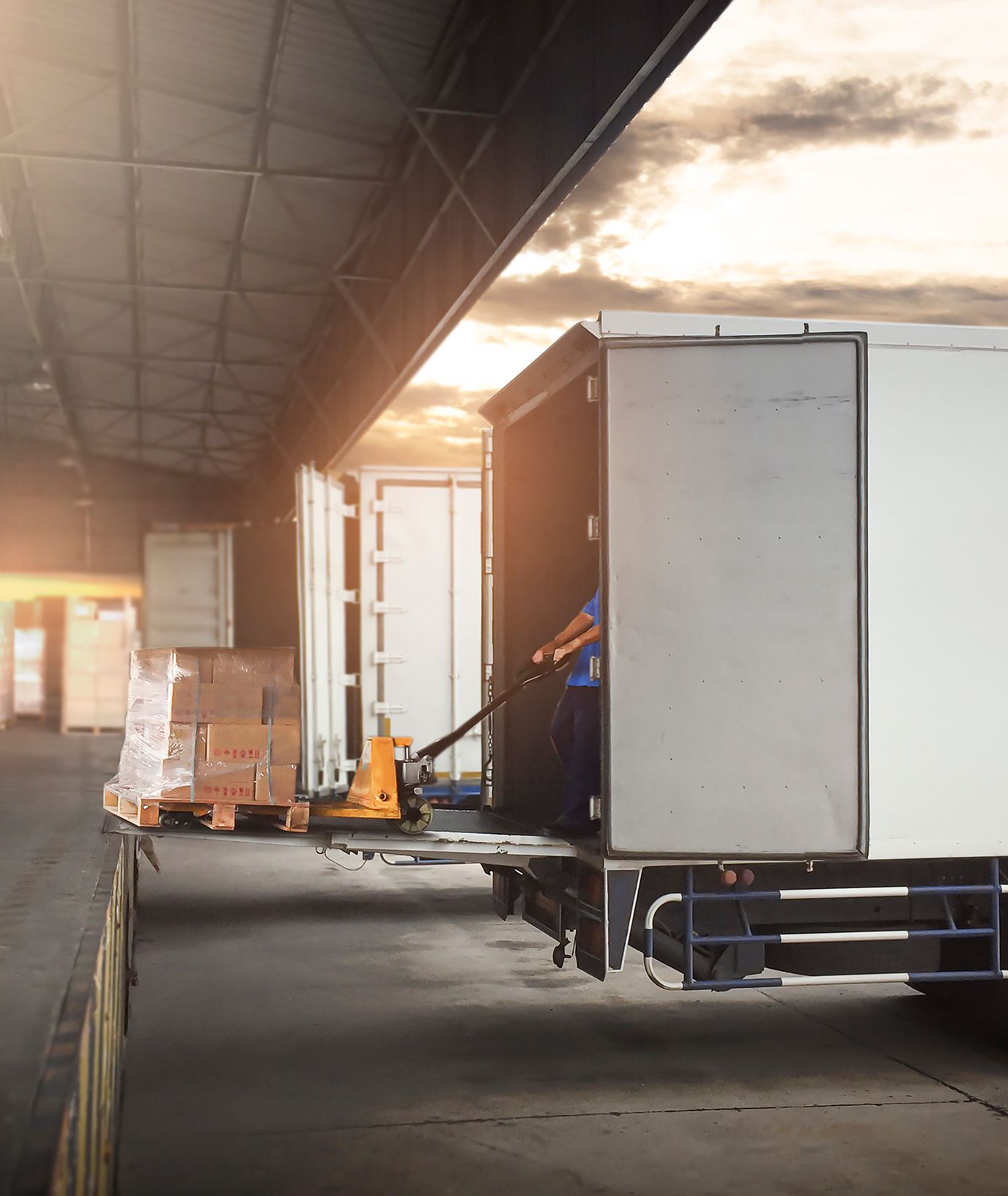 A worker uses a pallet jack to load or unload boxes from a truck at a warehouse dock as sunlight streams in from the background.