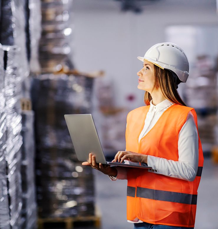 A woman wearing an orange safety vest and white hard hat uses a laptop while inspecting stacked pallets in a warehouse.