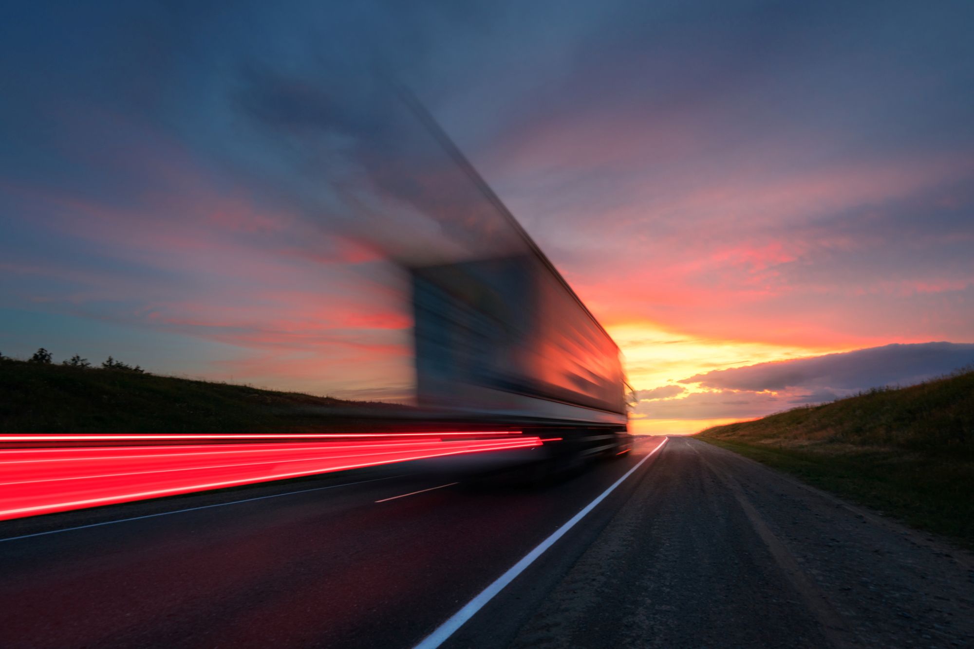 A blurred semi-truck speeds down a highway at sunset, creating red light trails against a colorful sky with clouds and glowing horizon.