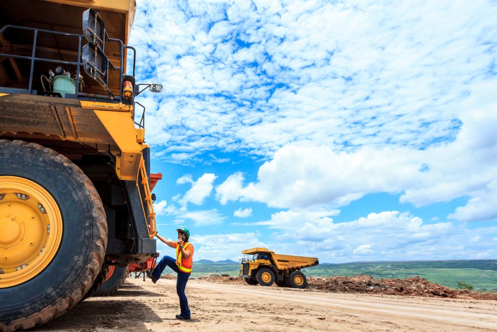 A worker in a safety vest and helmet climbs onto a large yellow mining truck on a dirt site, with another dump truck and blue sky with clouds in the background.