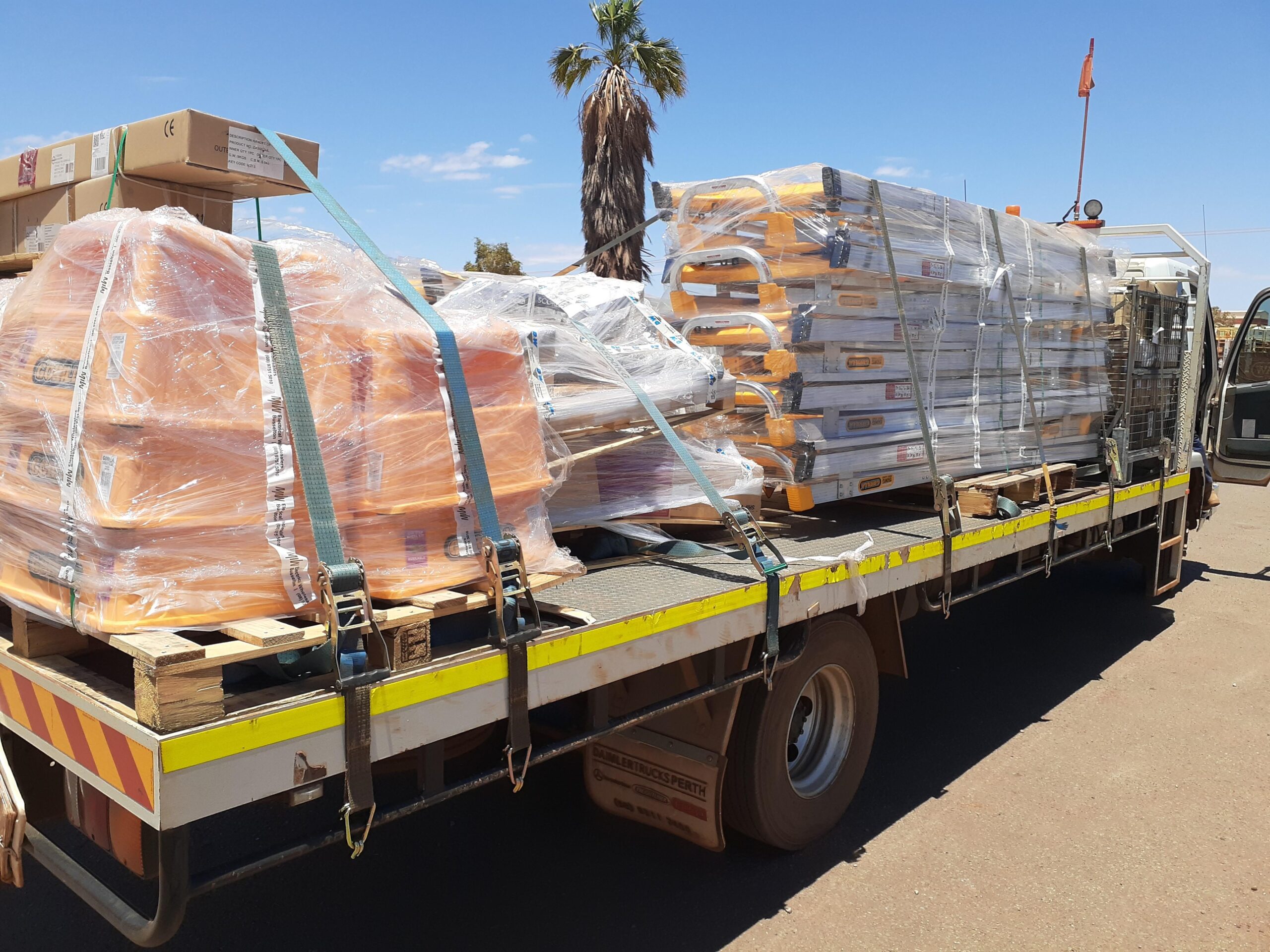 A flatbed truck loaded with stacks of large, orange plastic items and other packaged goods, all secured with plastic wrap and straps. A palm tree and blue sky are visible in the background.