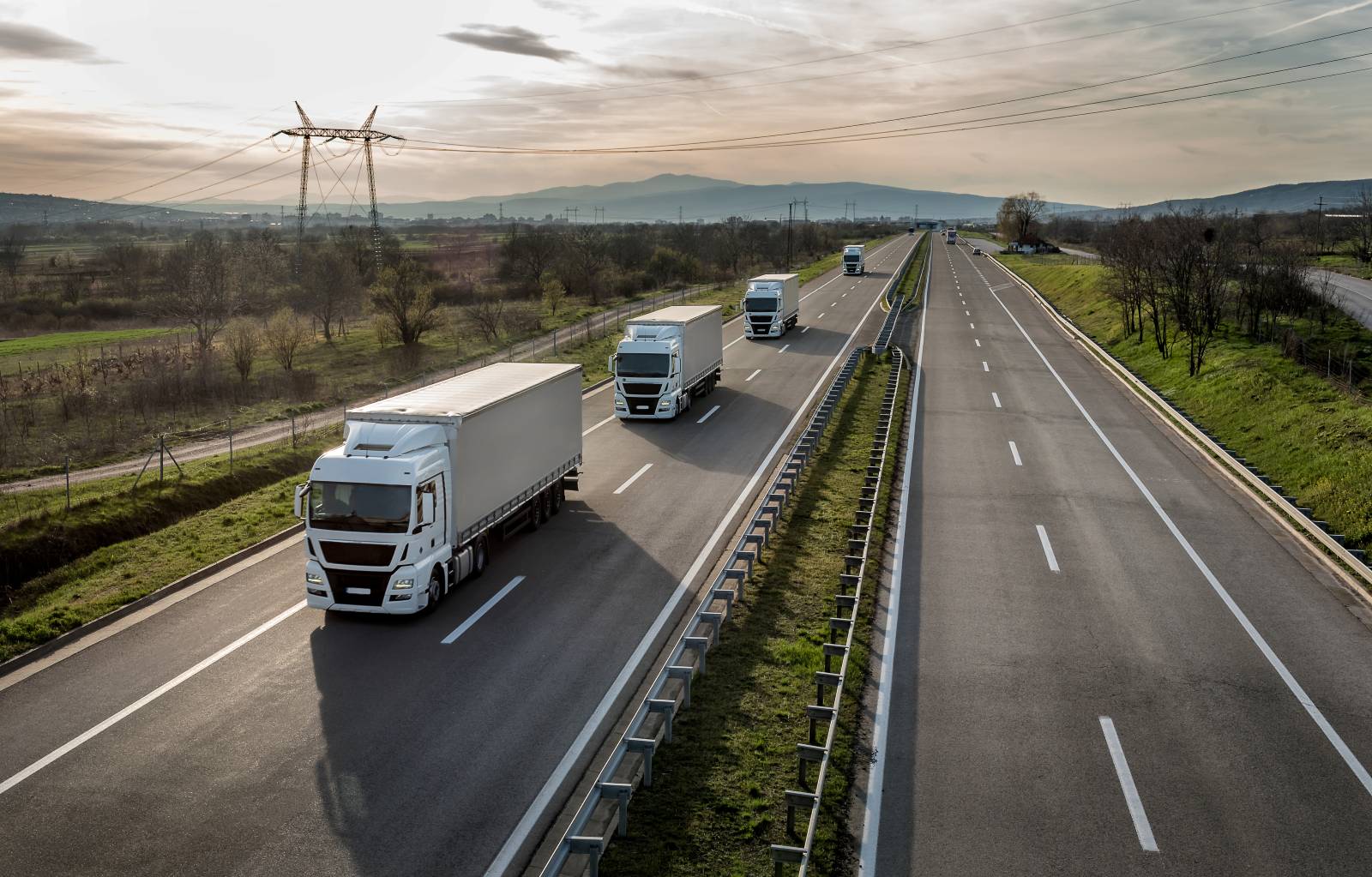 Several white semi-trucks drive along a divided highway through a rural landscape, with trees, fields, and distant hills under a cloudy sky. The road is mostly empty except for the trucks.