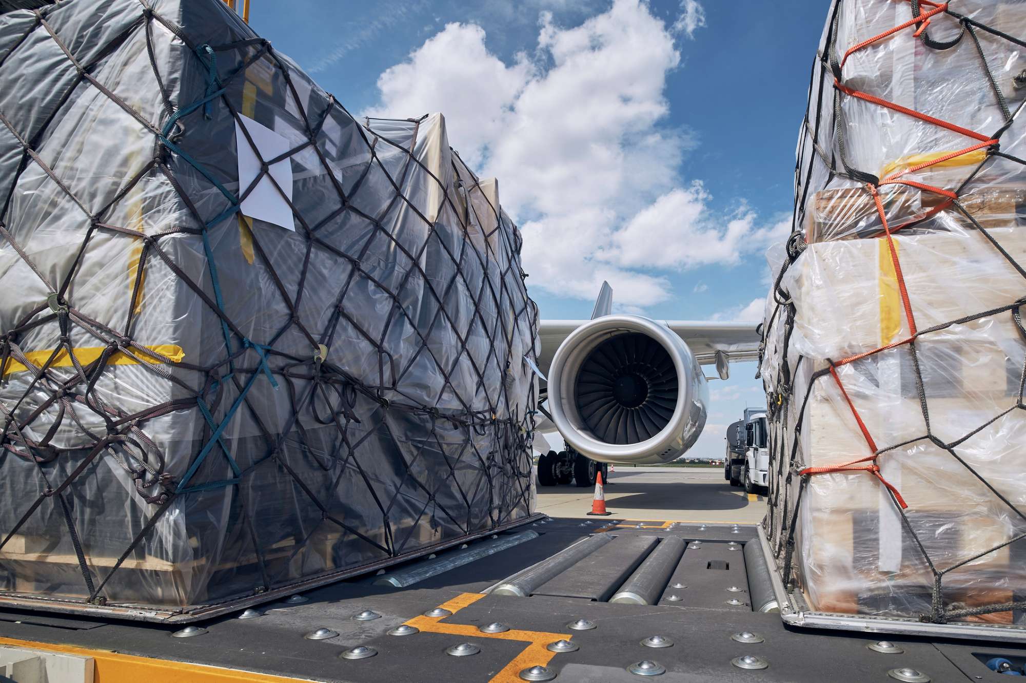 Large pallets of cargo wrapped in netting are loaded onto a plane, with the aircraft engine and landing gear visible in the background under a partly cloudy sky.