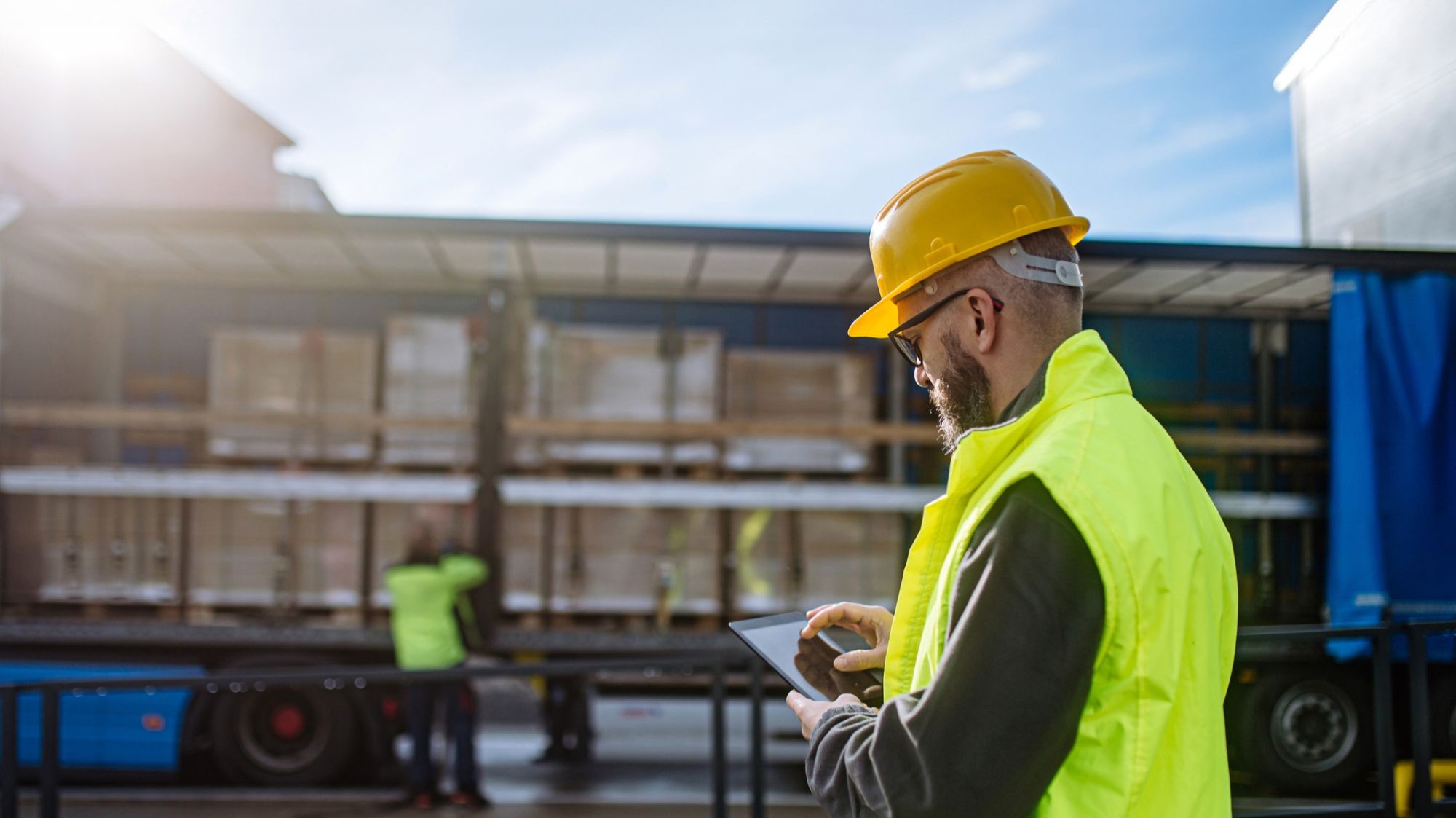 A worker wearing a yellow safety vest and hard hat uses a tablet near a truck loaded with pallets. Another worker is blurred in the background, unloading or organizing cargo. The scene is outdoors and sunny.