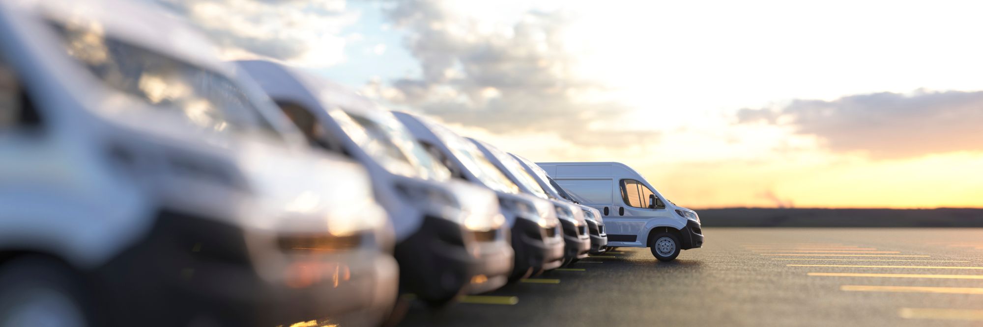 A row of white vans parked in a lot at sunset, with one van standing out in sharp focus while the others are blurred in the foreground. The sky is partly cloudy with a warm glow.