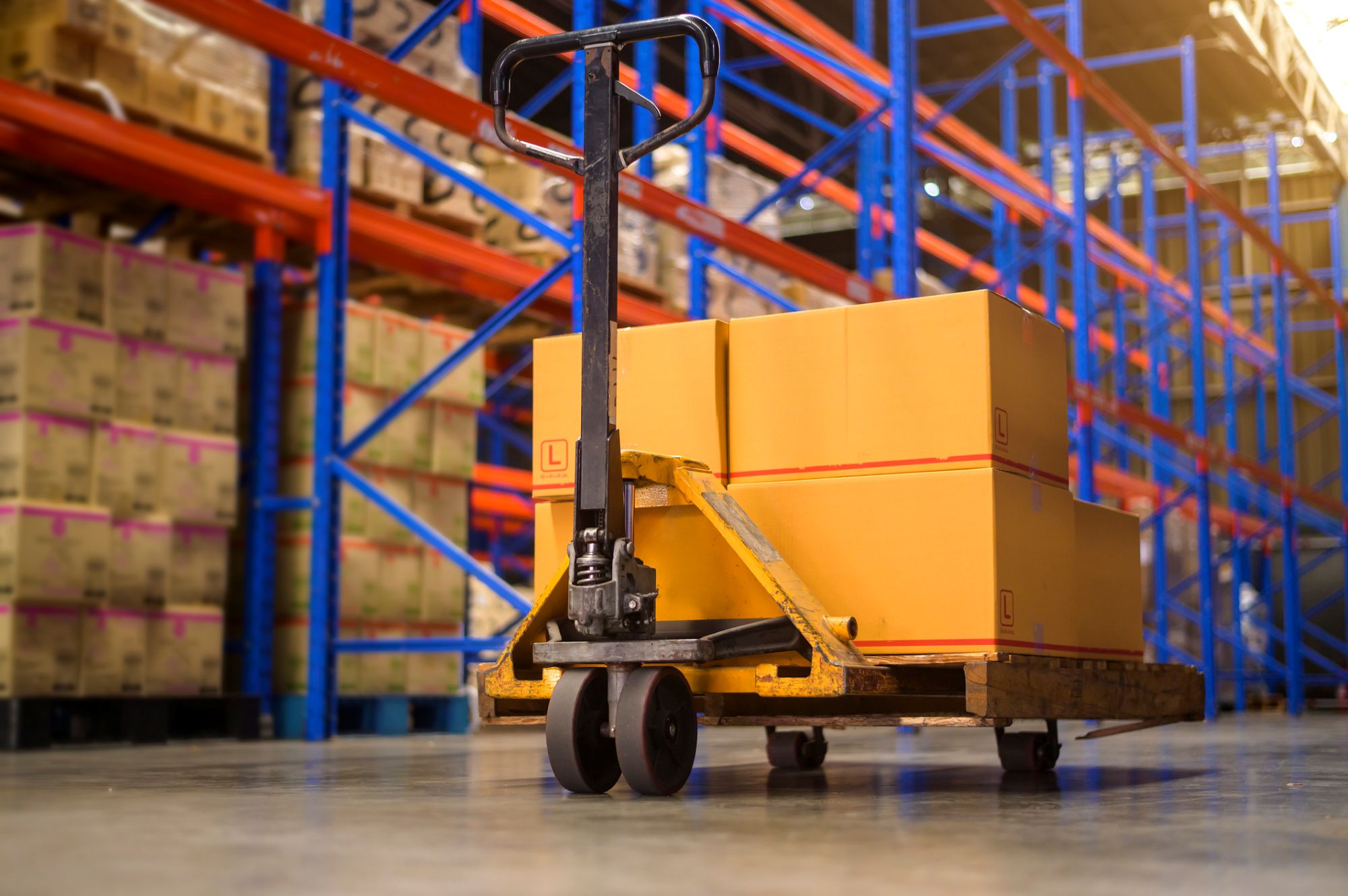 A pallet jack loaded with cardboard boxes sits on the floor of a warehouse, surrounded by tall blue and orange metal shelving filled with more boxes.