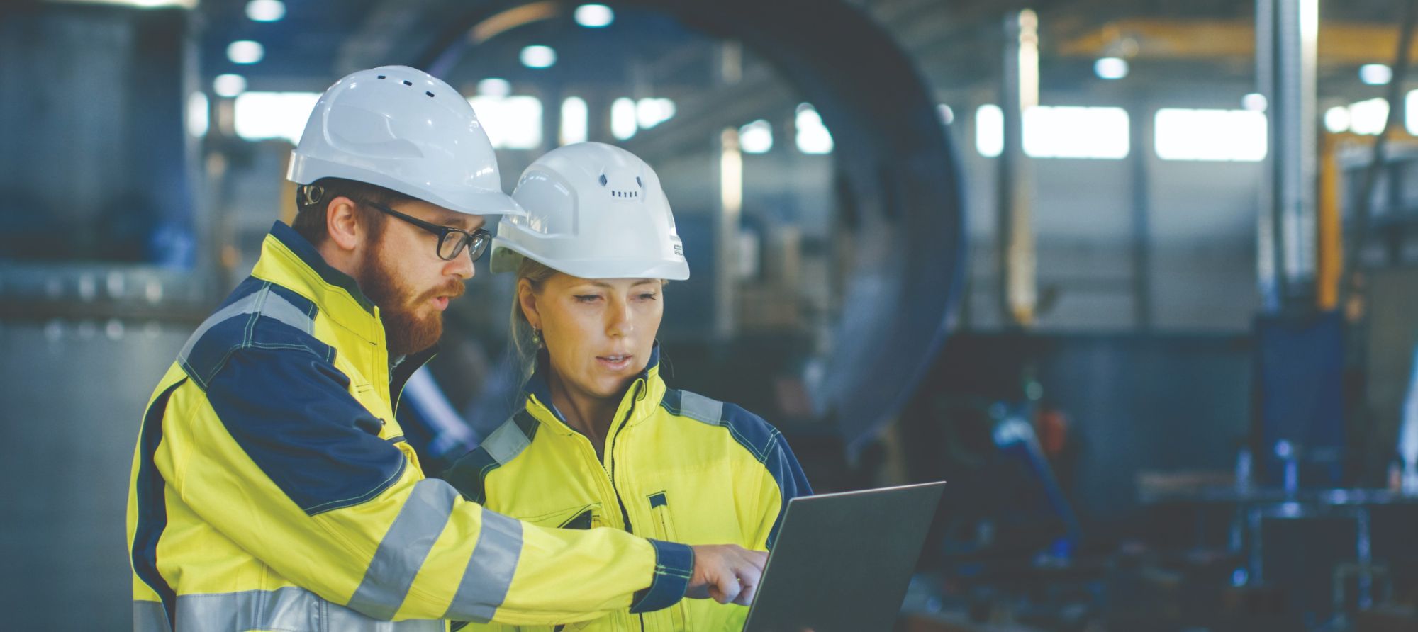 Two engineers wearing white helmets and yellow safety jackets stand in an industrial facility, looking at and discussing information on a laptop. Machinery and large equipment are visible in the background.